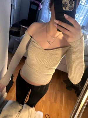 Glittrig offshoulder topp i beige - Trendig offshoulder topp i beige med glittrigt tyg och ribbad struktur. Långärmad och figurnära passform som framhäver axlarna 