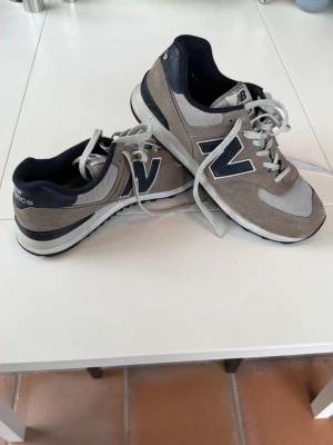 Grå New Balance sneakers - Snygga grå sneakers från New Balance med klassisk design. Skorna har mockadetaljer, meshpaneler och vita snören. Den ikoniska N-loggan syns tydligt på sidan och sulan är vit med svarta detaljer. Perfekta för en avslappnad streetstyle.