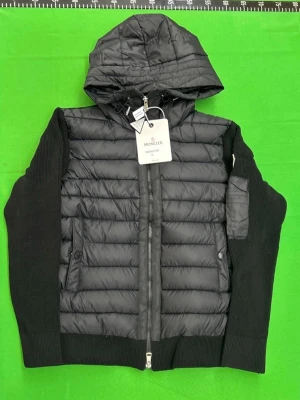 Svart dunväst med huva från Moncler - Snygg svart dunväst från Moncler med stickade svarta ärmar och vadderad kropp. Västen har en huva, dragkedja framtill, fickor med tryckknapp och en liten ficka med dragkedja på ärmen. Perfekt för dig som vill ha en stilren och modern look.