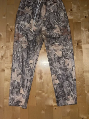 Camo mjukisbyxor från Realtree - Säljer ett par mjukisbyxor med camo-mönster från Realtree. Byxorna har elastisk midja med dragsko och muddar vid benslut. Materialet är mjukt och bekvämt, perfekt för chill dagar. Färgerna går i grått, beige och brunt med löv- och grenmotiv.