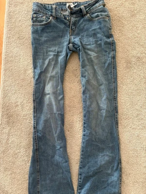 Blå bootcut jeans med slitningar - Säljer ett par blå bootcut jeans med klassiska fem fickor och snygga slitningar på framsidan. Jeansen har dragkedja och knapp framtill samt bälteshällor. Perfekt för dig som gillar en avslappnad och trendig look.
