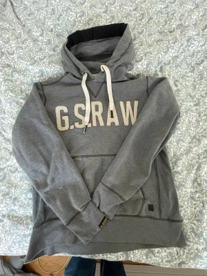 Grå hoodie från G-Star RAW - Säljer en grå hoodie från G-Star RAW med stor vit logotyp på bröstet och vita snören i huvan. Hoodien har en klassisk känguruficka och ribbade muddar. Materialet är mjukt och bekvämt, perfekt för chill dagar. Diskret svart logotypmärke på nederkanten.