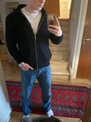 Svart cardigan med ribbad struktur - Säljer en svart cardigan med ribbad struktur och hög krage. Tröjan har dragkedja framtill och långa ärmar. Perfekt att slänga på över en t-shirt för en clean och enkel look.