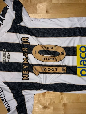 Santos FC Neymar Jr matchtröja - Säljer en svartvit randig Santos FC matchtröja från Umbro med Neymar Jr och nummer 10 på ryggen. Tröjan har flera sponsorloggor, rund hals och är tillverkad i lätt och ventilerande material. Perfekt för fotbollsfans som vill ha en ikonisk look.