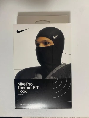 Nike Pro Therma-FIT Hood svart - Nike Pro Therma-FIT Hood är en svart balaclava med tight passform, perfekt för sport och träning. Tillverkad i ett syntetmaterial som håller dig varm och torr. Diskret Nike-logga på sidan och designad för att täcka hela huvudet.