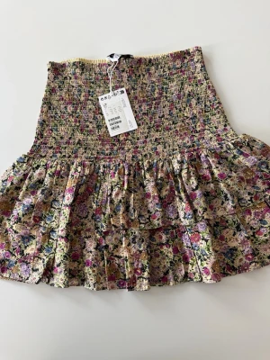 Blommig volangkjol från other stories  - Helt ny Supersöt kort blommig kjol från other stories med volang och smockad midja. Färgerna går i rosa, lila, grön och beige med små blommor över hela kjolen. Perfekt för dig som gillar romantisk och trendig stil.