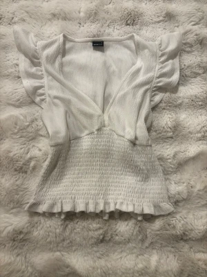 Vit blus med volang och v-ringning - Supersöt vit blus från Gina Tricot med v-ringning, volangdetaljer vid ärmslut och nederkant samt smockad midja. Toppen är perfekt att styla med jeans eller kjol.