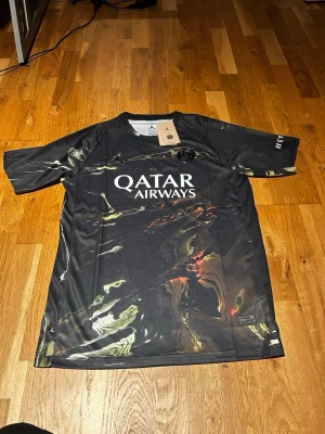 Psg 2025/26 away kit  - Cool svart PSG x Jordan matchtröja med unikt mönster i guld och silver. Framsidan har Qatar Airways-logga och PSG-emblem, medan ärmen har texten 'BEND'. Tillverkad i lätt och ventilerande material, perfekt för fotboll eller träning.