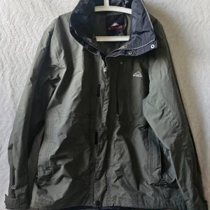 Grön vindjacka från McKinley - Snygg grön vindjacka från McKinley med hög krage och dragkedja framtill. Jackan har två sidofickor med dragkedja och justerbar nederkant. Materialet är slitstarkt och vattenavvisande, perfekt för blåsiga dagar. Logotyp på bröstet och svart innerfoder.