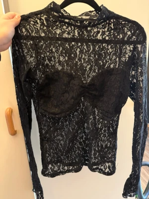Svart spetsblus med hög krage - Snygg svart långärmad spetsblus med hög krage och transparent material. Toppen har en markerad byst med satindetalj och vacker spets över hela plagget. Perfekt för dig som vill ha en edgy och trendig look.