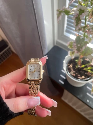 Guldfärgad klocka  - Snygg guldfärgad klocka  med rektangulärt ur och vit urtavla. Klockan har en länkad metallrem och minimalistisk design med guldiga visare och index. Perfekt accessoar för dig som gillar stilrena detaljer.