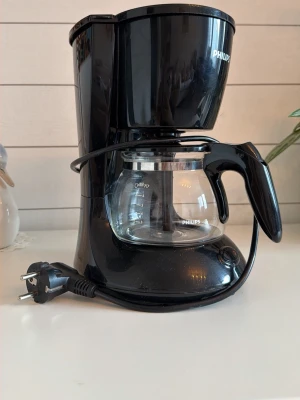 Svart kaffebryggare Philips HD7432 - Säljer en svart kaffebryggare från Philips, modell HD7432. Bryggaren har en glaskanna med svart handtag och tydliga markeringar för vattennivå. Strömkabeln är fastmonterad och maskinen har en kompakt design som passar i de flesta kök.