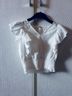 Vit blus med volang och broderi - Söt vit blus med v-ringning, volangdetaljer på axlarna och broderade mönster. Smockad midja ger en snygg passform och toppen är ärmlös. Perfekt för dig som gillar romantisk och feminin stil.