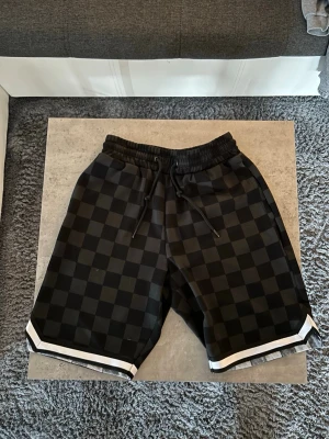 Svarta rutiga shorts med snörning - Säljer ett par svarta shorts med rutigt mönster och vita detaljer längst ner. Shortsen har elastisk midja med snörning och fickor både fram och bak. Perfekta för en avslappnad och stilren look.