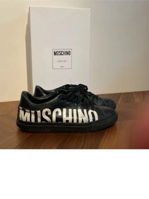 Svarta sneakers från Moschino - Coola svarta sneakers från Moschino med stort vitt logotryck på sidan. Skorna har klassisk snörning och är tillverkade i skinn med en låg profil och svart sula. Perfekta för dig som vill sticka ut med ett statement-märke.