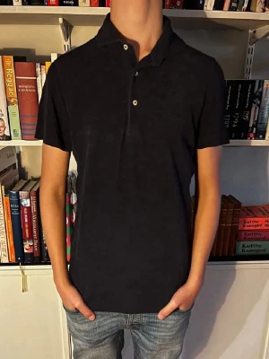 Mörkblå frottépiké med kort ärm - En enkel och stilren svart polotopp med korta ärmar och knappar framtill. Perfekt för dig som gillar en klassisk look. Materialet känns mjukt och bekvämt, troligen bomull eller polyester. Passformen är normal och toppen har en snygg krage.