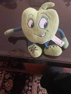 Grön hjärtformad mjukisfigur - Säljer en grön hjärtformad mjukisfigur med broderade ögon och mun, samt armar och ben i grönt och blått. Figuren är tillverkad i mjukt plyschmaterial och har en lekfull design med ett litet blad på toppen.