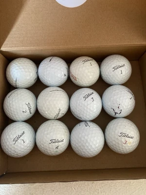 Golfbollar Titleist i kartong - Säljer ett set med 12 vita golfbollar från Titleist i A/B klass,  med märkning som Pro V1 och olika siffror. Bollarna har klassisk dimple-design och är tillverkade i slitstarkt material. Perfekt för dig som vill fylla på golfutrustningen.