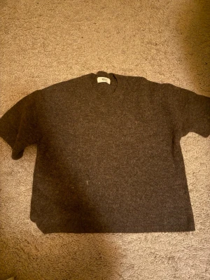 Mörkbrun stickad tröja - En enkel och stilren mörkbrun stickad tröja med rund hals och korta ärmar. Tröjan har en rak passform och är gjord i ett mjukt, tjockt material som passar perfekt för kyligare dagar.
