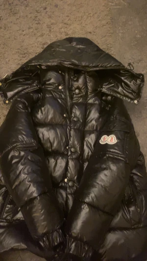 Svart Moncler dunjacka med huva - Säljer en svart, glansig dunjacka.   Moncler med stor huva och klassisk logga på ärmen. Jackan har quiltad design, dragkedja och knappar framtill samt elastiska muddar. Perfekt för kalla dagar när du vill vara både varm och stilren.                                                            Passar dig som är från 165-170, pris kan diskuteras kommer med tags och authenticate