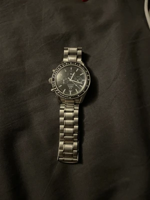 Omega Speedmaster kronograf klocka - Klassisk Omega Speedmaster kronograf med svart urtavla och silverfärgad länk i rostfritt stål. Klockan är i nytt skick bara att själv bandet har lossnat men biten får man med vid köp. Annars är de bara köpa en ny så blir klockan som ny