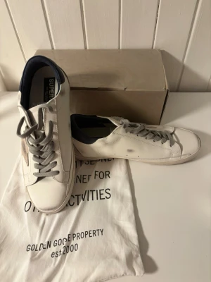 Vita Golden Goose sneakers - Snygga vita sneakers från Golden Goose med grå skosnören och mörkblå detaljer vid hälen. Klassisk låg modell i läder med diskret stjärndetalj på sidan. Kommer med originalpåse och låda. Perfekta för dig som gillar stilrena och trendiga skor.