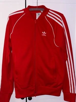 Röd Adidas track jacket med vita ränder - Säljer en klassisk röd Adidas track jacket med dragkedja, vita ränder längs ärmarna och vit piping. Tröjan har två fickor fram och Adidas logga på bröstet. Perfekt för en sportig och retro look.