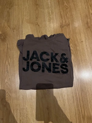 Brun t-shirt från Jack & Jones - Snygg brun t-shirt från Jack & Jones med stor svart logga framtill. Klassisk rund hals och korta ärmar. Tillverkad i mjuk bomull som känns skön mot huden. Perfekt för en avslappnad och trendig stil.