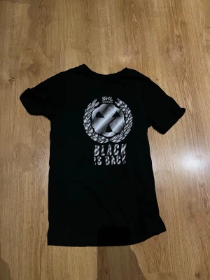 Svart t-shirt Black is Back - Svart t-shirt med tryck i silver fram på tröjan där det står 'Black is Back' och ett stort emblem. T-shirten har rund hals och korta ärmar, perfekt för en cool och avslappnad stil. En riktig snygg Brynäs      t-shirt.