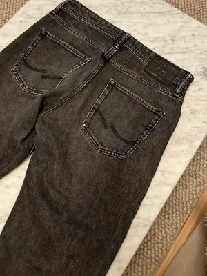 Svarta jeans från Jack & Jones - Säljer ett par svarta jeans från Jack & Jones med klassisk femficksmodell och raka ben. Jeansen har en tvättad look och tydliga sömmar, samt svart knapp och dragkedja. Perfekta för en avslappnad och trendig stil.