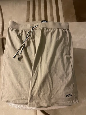 Beige shorts från BOSS Homewear - Snygga beige shorts från BOSS Homewear med elastisk midja och dragsko i svart och vitt. Tillverkade i mjukt material, perfekta för en avslappnad stil. Diskret BOSS-logga nertill på benet och praktiska sidofickor. Man kan köpa (komplett sett) skriv för mer info 