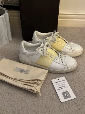 Äkta Valentinos - Snygga vita sneakers från Valentino med en bred gul panel på sidan och vita skosnören. Skorna har en klassisk låg siluett och är tillverkade i slätt läder. Perfekta för dig som vill ha en stilren men ändå unik look. Skorna har en liten röd fläck men den syns inte vid användning