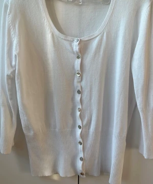 Vit ribbad kofta med knappar - Stilren vit kofta med långa ärmar och ribbade muddar. Koftan har rund halsringning och knäpps med silvriga knappar framtill. Perfekt att slänga över en topp för en enkel och fräsch look.