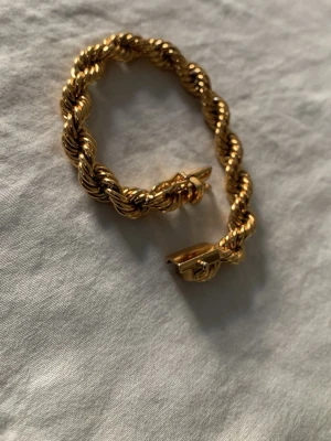 Tvinnat guldarmband i metall - Säljer ett chunky armband i guldfärgad metall med tvinnad design. Armbandet har ett klassiskt lås och känns riktigt statement med sin grova kedja. Perfekt för dig som gillar att sticka ut med accessoarerna och vill ha något som syns.