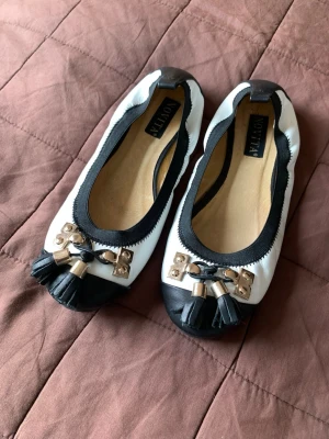 Ballerinaskor - Novita - Svart/vita ballerinaskor med gulddetaljer från Novita. Storlek 36. Lite sliten längst fram vid tån, se två sista bilderna. I övrigt i fint skick 🌸