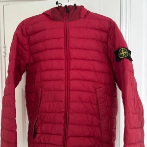 Röd dunjacka från Stone Island - Vinröd stone island dun jacka med huva. Den ser rödare ut på bilderna men skulle säga att den är mörkare röd. Flera år gamal men i väldigt fint skick. Perfekt nu till våren.