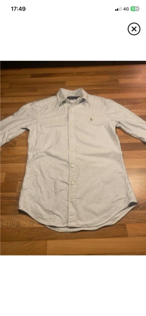 Randig skjorta från Ralph Lauren - Klassisk vit skjorta med tunna blå ränder från Ralph Lauren. Skjortan har lång ärm, knappar framtill och den ikoniska broderade loggan på bröstet. Tillverkad i bomull för en skön och luftig känsla.