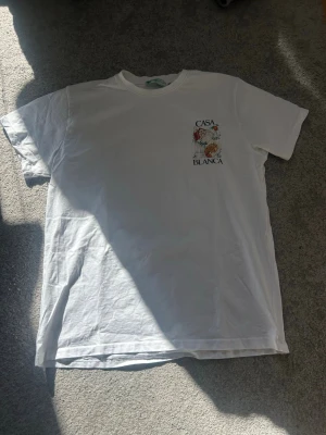 Vit Casablanca t-shirt - Säljer en vit t-shirt från Casablanca Tennis Club med ett litet färgglatt tryck och texten 'Casa Blanca' på bröstet. Klassisk passform och rund hals. Tillverkad i mjuk bomull som känns skön mot huden. Perfekt för dig som gillar stilrena plagg med en twist.