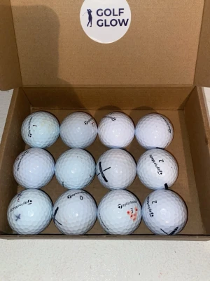 Taylormade mix 12-pack - Säljer ett set med 12 vita golfbollar från TaylorMade i en brun kartong med Golf Glow-klistermärke. Bollarna har olika markeringar och siffror, perfekta för dig som vill fylla på golfväskan.