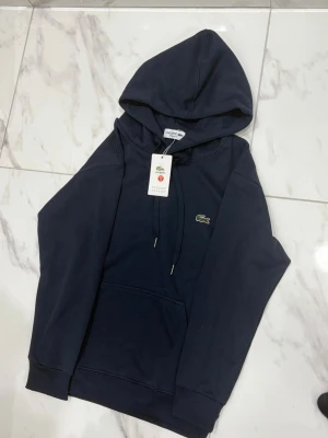 Mörkblå hoodie från Lacoste - Snygg mörkblå hoodie från Lacoste med klassisk krokodillogga på bröstet. Regular fit med huva och dragsko, samt en stor magficka framtill. Tillverkad i mjukt bomullsmaterial som känns skönt mot huden. Perfekt för en avslappnad och stilren look.