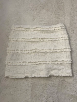 Vit spetskjol med volangdetaljer  - Supersöt vit kortkjol med flera lager volang och spetsdetaljer. Kjolen har en feminin och romantisk look med vackert spetsmönster och volanger som ger extra struktur. Perfekt för dig som vill ha en söt och trendig kjol till sommaren. minikjol 