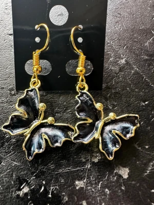 Svarta fjärilsörhängen med guldkant - Unika örhängen i form av fjärilar med svart emalj och guldfärgade kanter. Hängande modell med krok, perfekta för dig som gillar statement-accessoarer. Materialet är metall med emaljerad yta. Säljs på originalkortet från Fashion Jewelry.