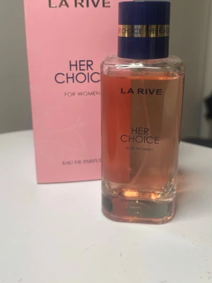 La Rive Her Choice Eau de Parfum - La Rive Her Choice Eau de Parfum för kvinnor i en elegant, rektangulär glasflaska med rosa doftvätska och blått lock med gulddetaljer. Kommer i en rosa kartong med stilren design. Perfekt för dig som gillar feminina och fräscha dofter.