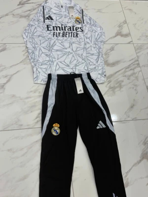 Real Madrid träningsset Adidas - Snyggt träningsset från Adidas med Real Madrid-logga. Överdelen är vit med grått grafiskt mönster, svart Adidas-logga och 'Emirates Fly Better'-tryck. Byxorna är svarta med vita detaljer och klubbmärke. Perfekt för fotbollsträning eller chill.