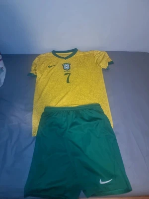 Brasilien matchställ Nike Vini Jr - Säljer ett Brasilien matchställ från Nike med gul tröja och gröna shorts. Tröjan har nummer 7 och Vini Jr på ryggen samt landslagets emblem på bröstet. Setet är gjort i lätt och ventilerande material, perfekt för fotbollsträning eller match.