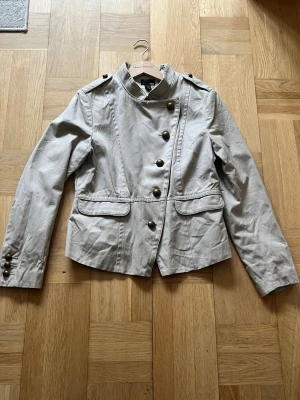 Napoleon jacka - Snygg beige rock från h&m med dekorativa metallknappar i dubbla rader, axelklaffar och ståkrage. Jackan har två fickor framtill och lång ärm med knappar vid ärmslut. Perfekt för dig som gillar en stilren och trendig look. Kom gärna med prisförslag