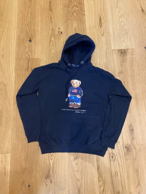 Marinblå hoodie Polo Bear Ralph Lauren - Snygg marinblå hoodie från Ralph Lauren med den ikoniska Polo Bear-trycket på bröstet. Hoodien har huva med dragsko, stor magficka och ribbade muddar. Tillverkad i mjukt bomullsmaterial som är perfekt för en avslappnad och trendig stil.