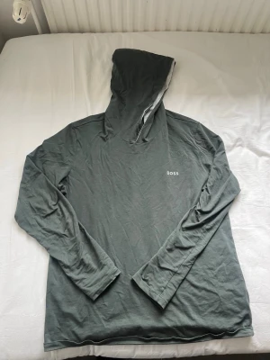 Grön hoodie från BOSS med huva - Säljer en stilren grön hoodie från BOSS i storlek S. Hoodien har en klassisk huva och lång ärm, samt en diskret BOSS-logga på bröstet. Tillverkad i mjukt och tunt material som känns skönt mot huden. Perfekt för en avslappnad look.