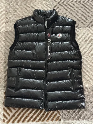 Svart dunväst från Moncler - Snygg svart dunväst från Moncler med glansig finish och broderad logga på bröstet. Västen har hög krage, två dragkedjeförsedda fickor och tydlig Moncler-detalj vid dragkedjan. Perfekt för lager-på-lager och streetstyle.
