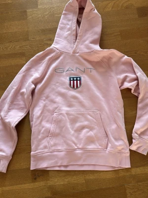 Rosa hoodie från GANT med logga - Snygg rosa hoodie från GANT med broderad logga och sköld på bröstet. Klassisk känguruficka framtill och stor huva. Tillverkad i mjukt bomullsmaterial som är skönt att ha på sig. Perfekt för dig som gillar en avslappnad och sportig stil.